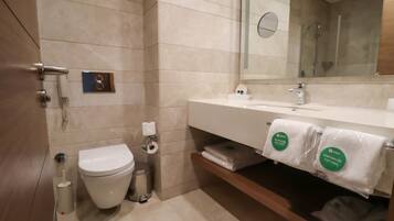 A modern bathroom with a toilet, sink, and mirror. There are towels and a trash can visible.