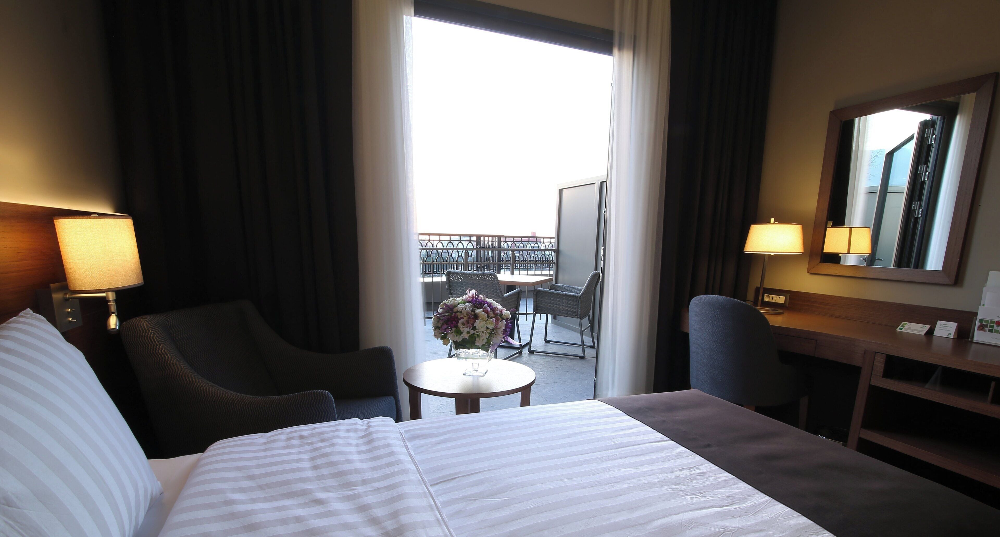 Een hotelkamer met een bed, twee stoelen, een kleine tafel met een vaas, een bureau met een lamp, en een balkon met buitenmeubels.