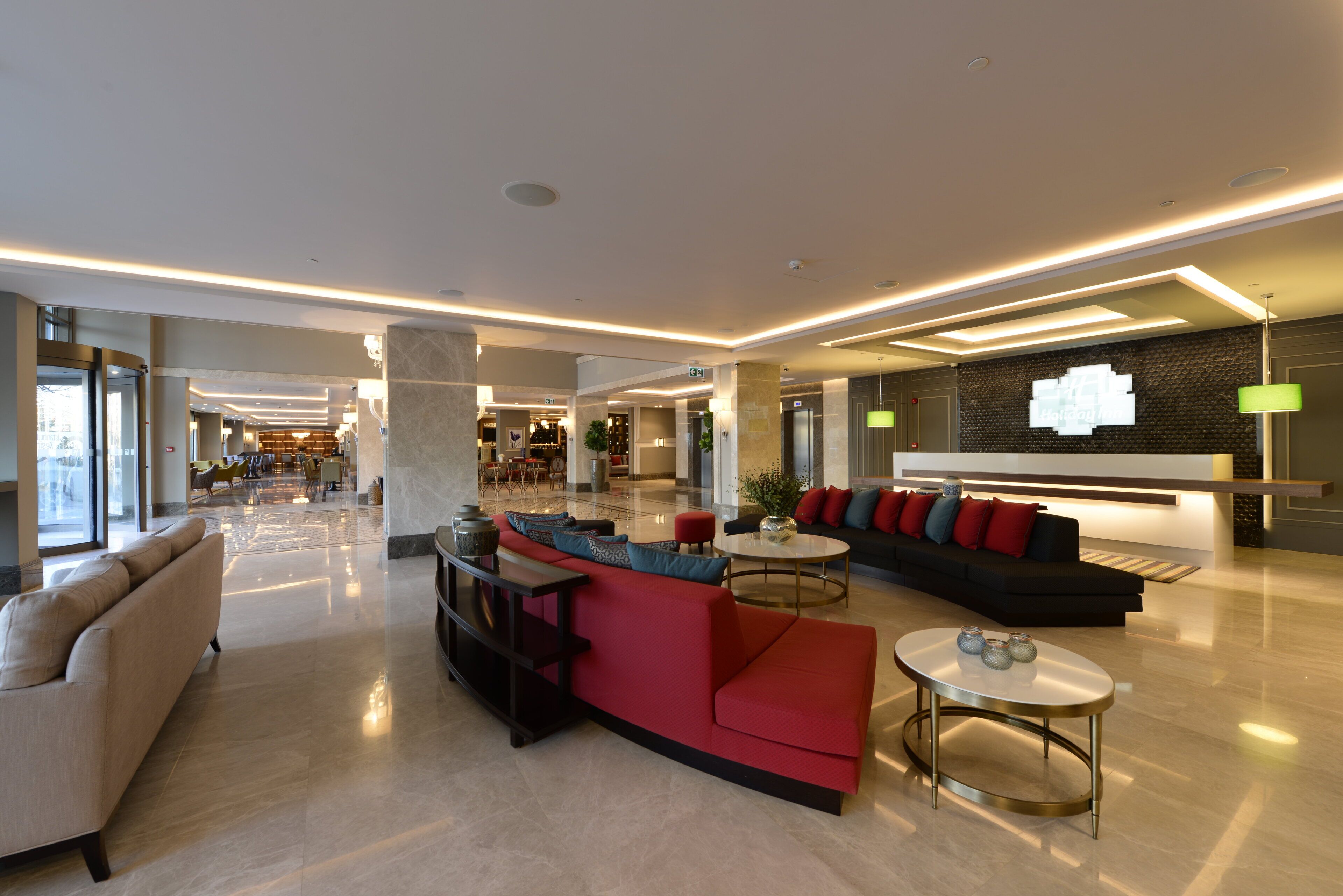 A spacious lobby with a red sofa, beige armchairs, and a black couch. There are multiple tables and a reception desk with a sign.