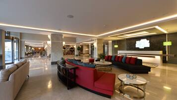 Eine geräumige Lobby mit einem roten Sofa, beigen Sesseln und einem schwarzen Couchsofa. Es gibt mehrere Tische und eine Rezeption mit einem Schild.