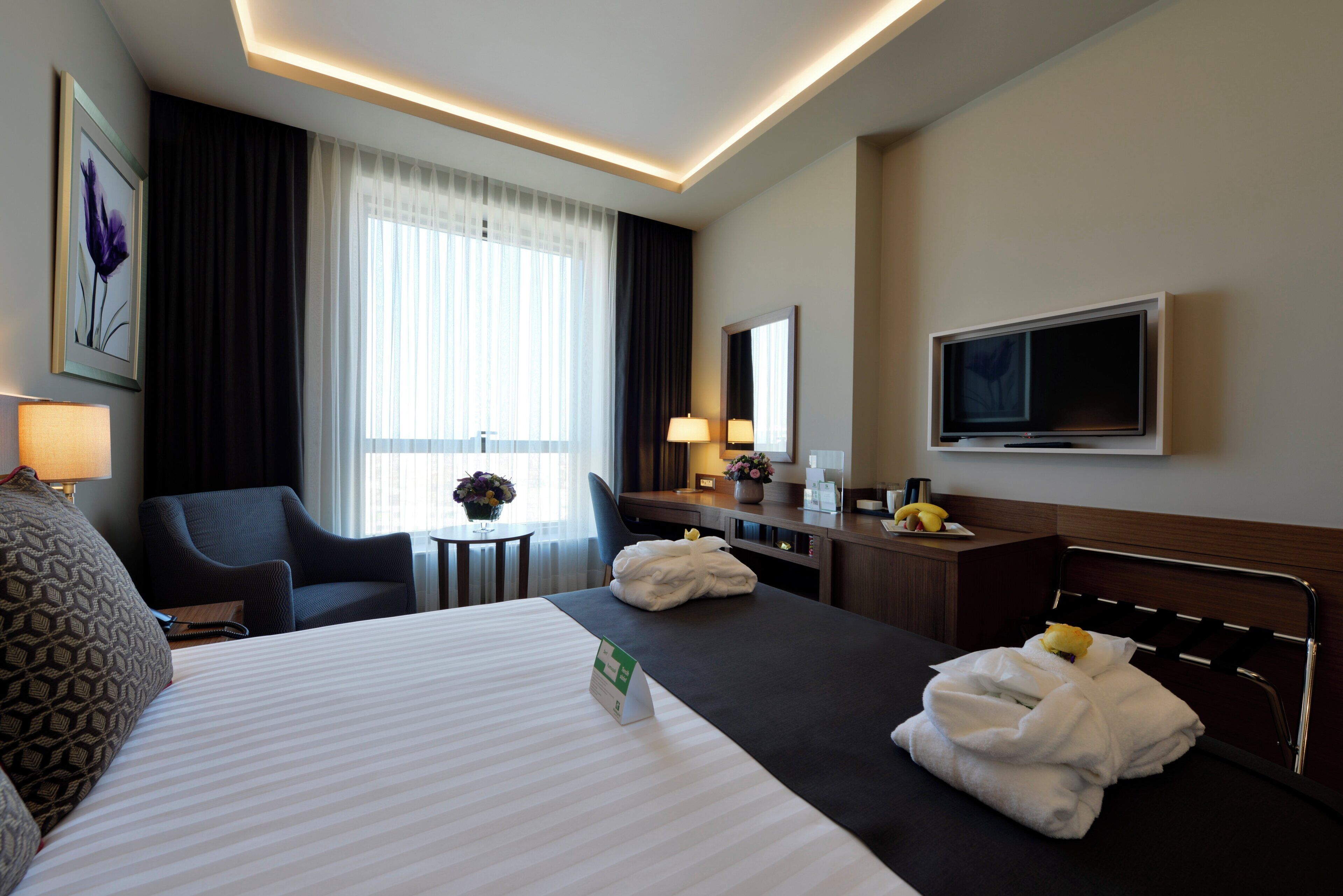 A hotel room with a bed, two chairs, a desk, a television, and a window with curtains.