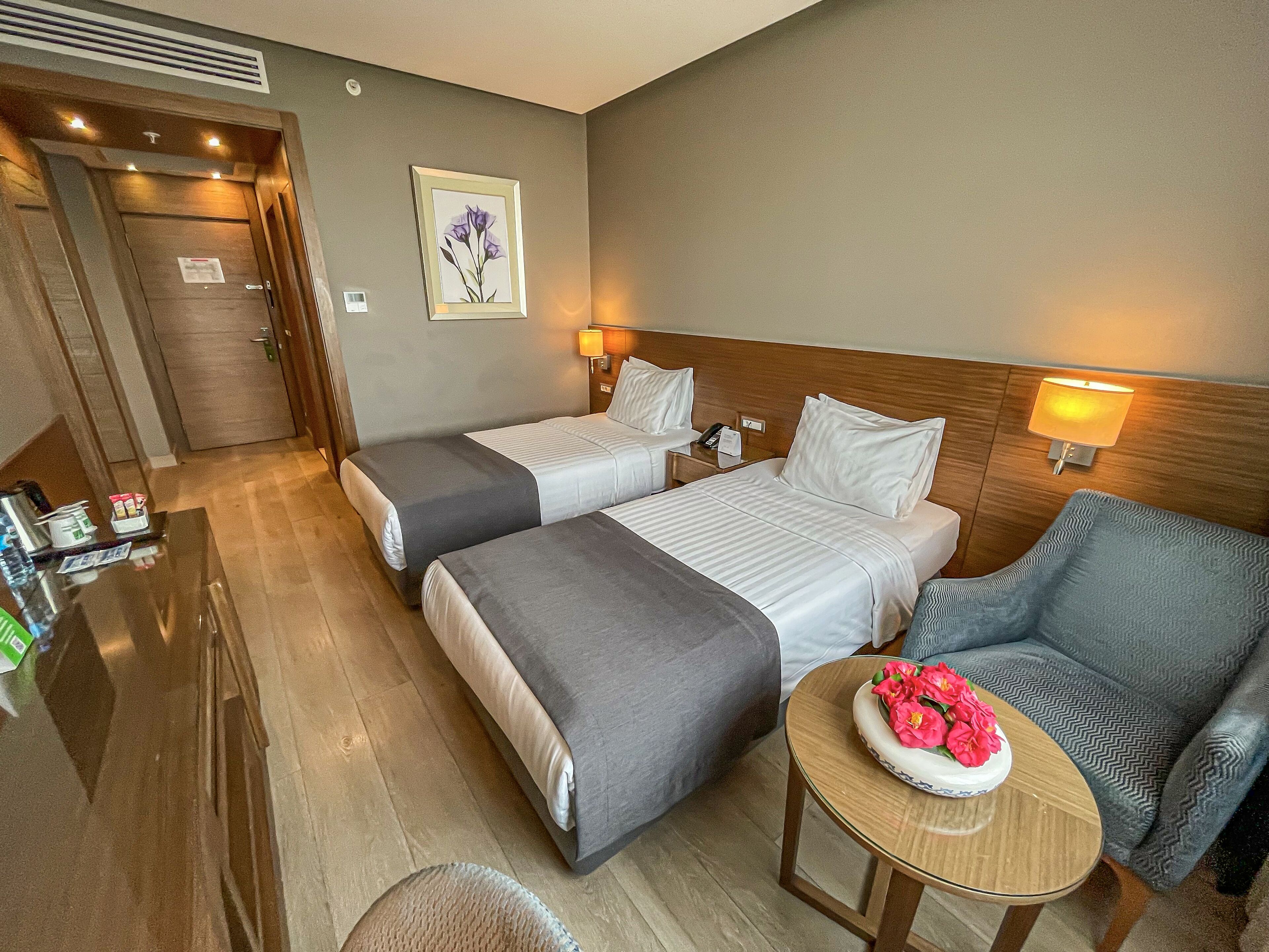 A hotel room with two beds, a desk, a chair, and a small table with flowers.