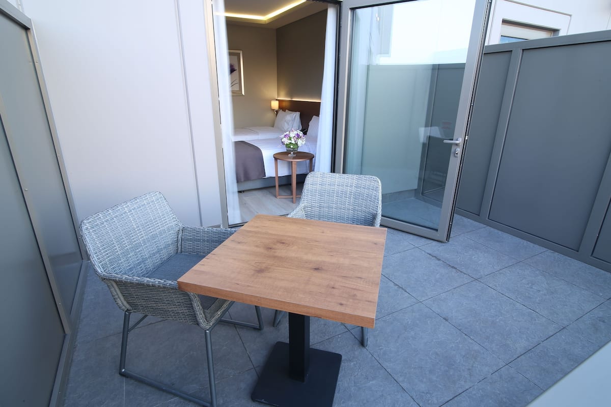 Ein Balkon mit einem Holztisch und Korbstühlen, ein Bett mit weißem Kopfteil sowie ein kleiner runder Tisch mit einer Blumenvase.
