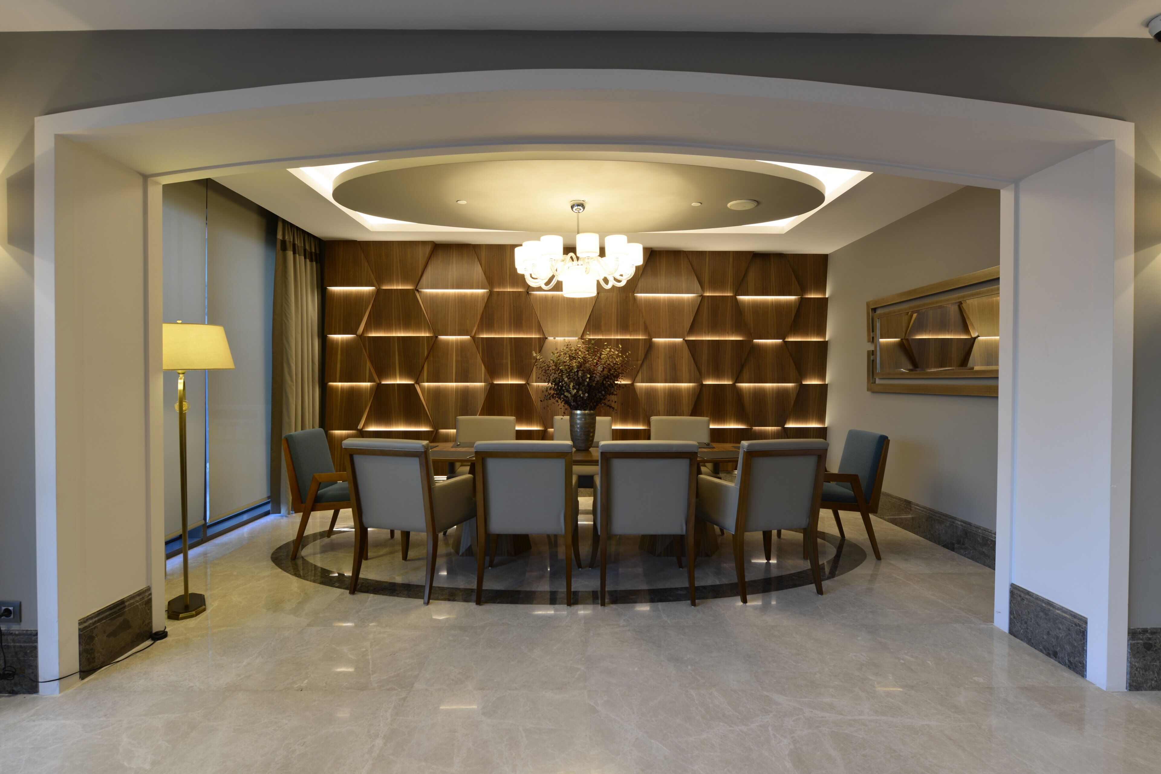 A modern dining area with a round table, chairs, a chandelier, and a decorative wall panel.