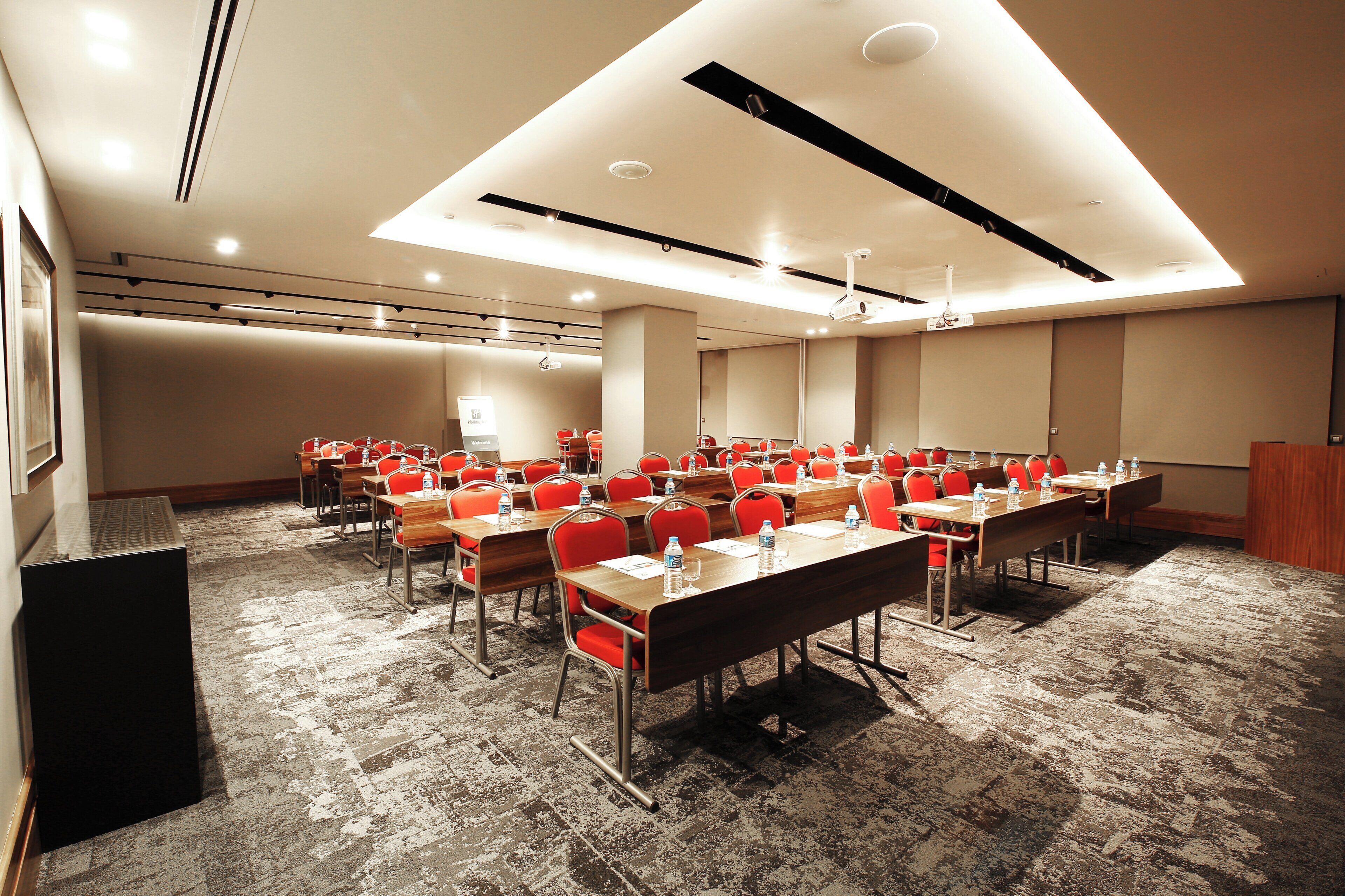 A conference room with multiple tables and chairs, equipped with water bottles and papers, and a projector mounted on the ceiling.