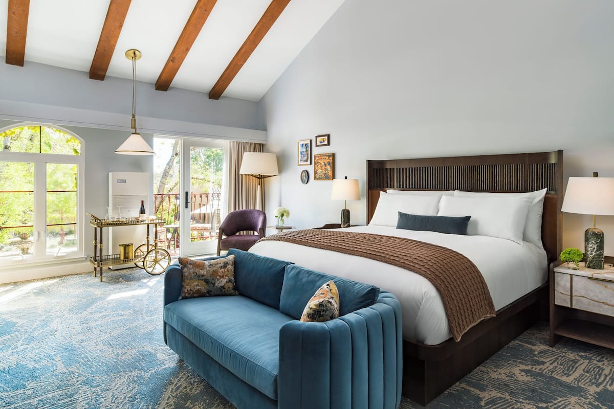 A modern hotel room with a large bed, a blue sofa, a dining area with a table and chairs, and a balcony with a view of trees.