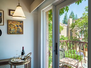 A room with a dining table, a wine bottle, and a chair. There is a window with a view of greenery and buildings outside.