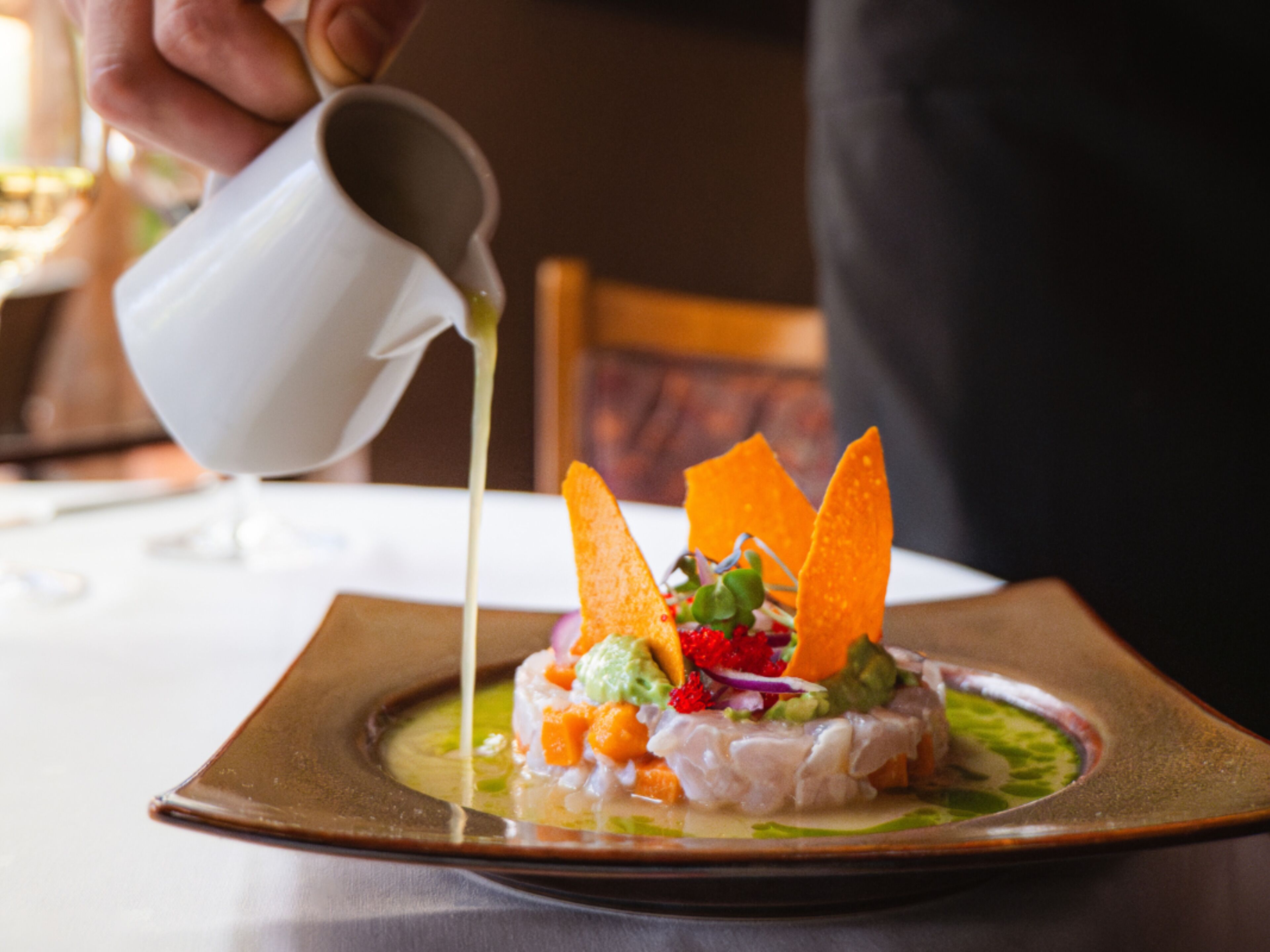 A dish with a square plate, a white pitcher pouring a clear liquid, and a person in dark clothing in the background.