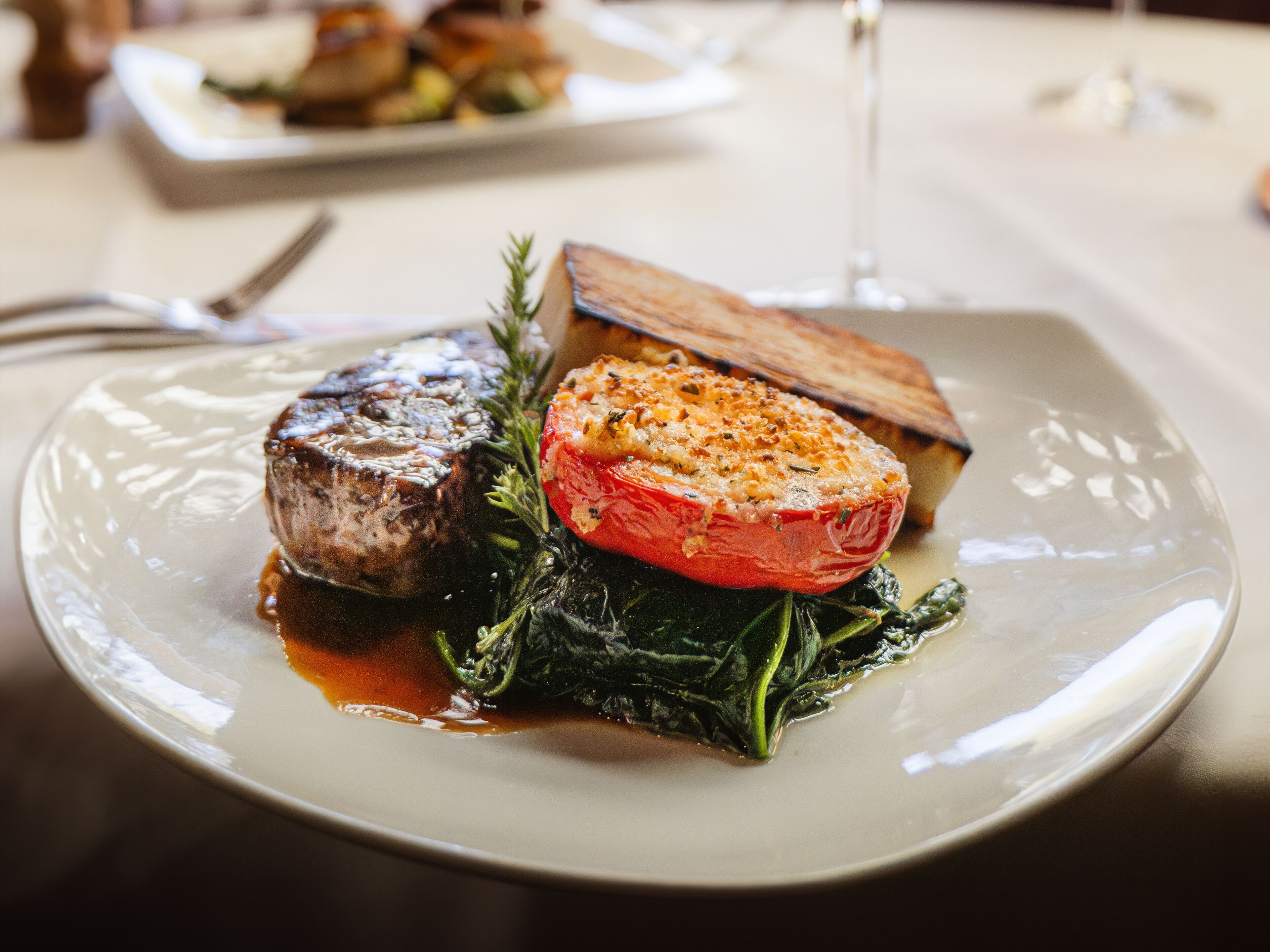 Un plato de comida con bistec, tomate asado y verduras de hoja verde.