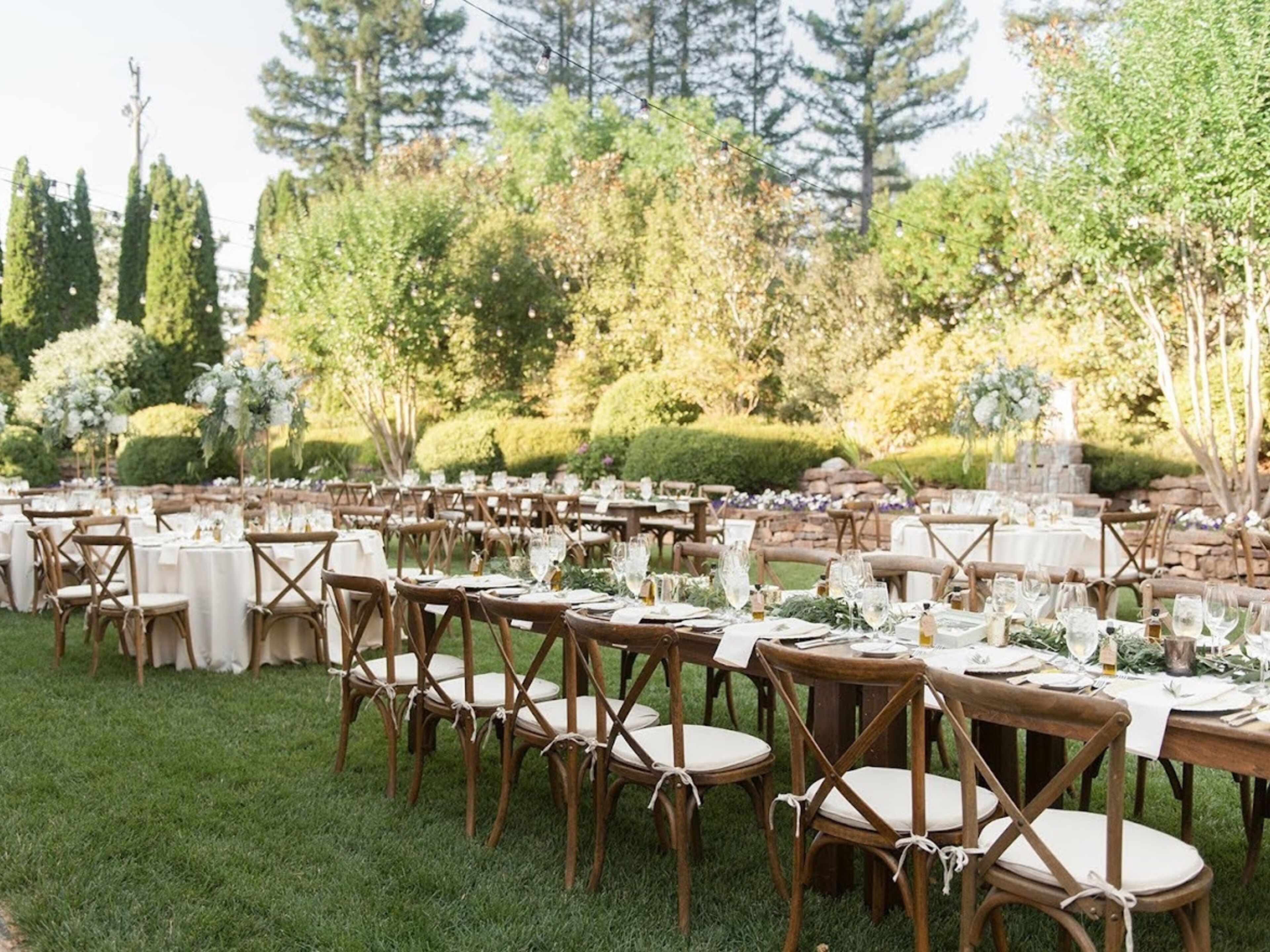 An outdoor dining area with round tables and chairs, surrounded by greenery and trees.