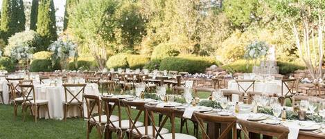 An outdoor dining area with round tables and chairs, surrounded by greenery and trees.