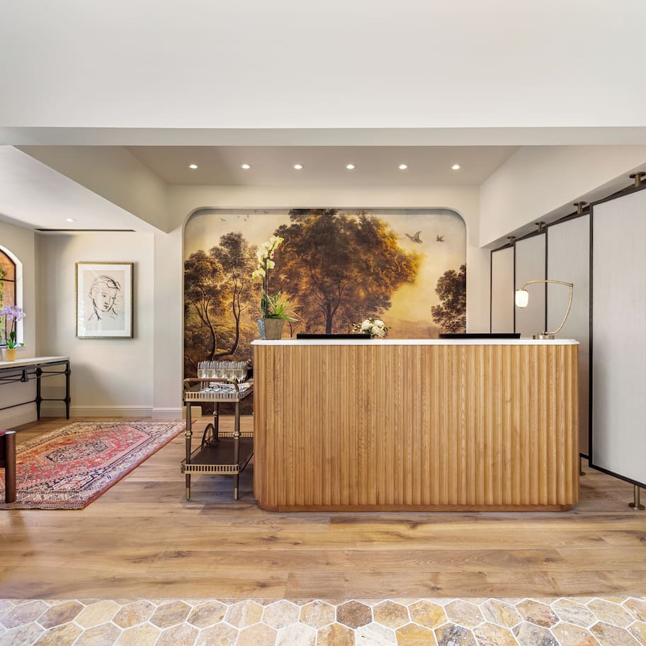 A reception area with a wooden reception desk, a stone-tiled floor, a red chair, a patterned rug, and a mural of trees.