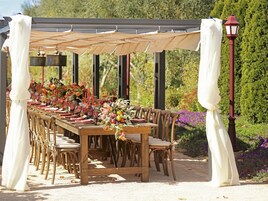 Un área de comedor al aire libre con una larga mesa de madera dispuesta para una comida, rodeada de sillas y una pérgola en la parte superior.