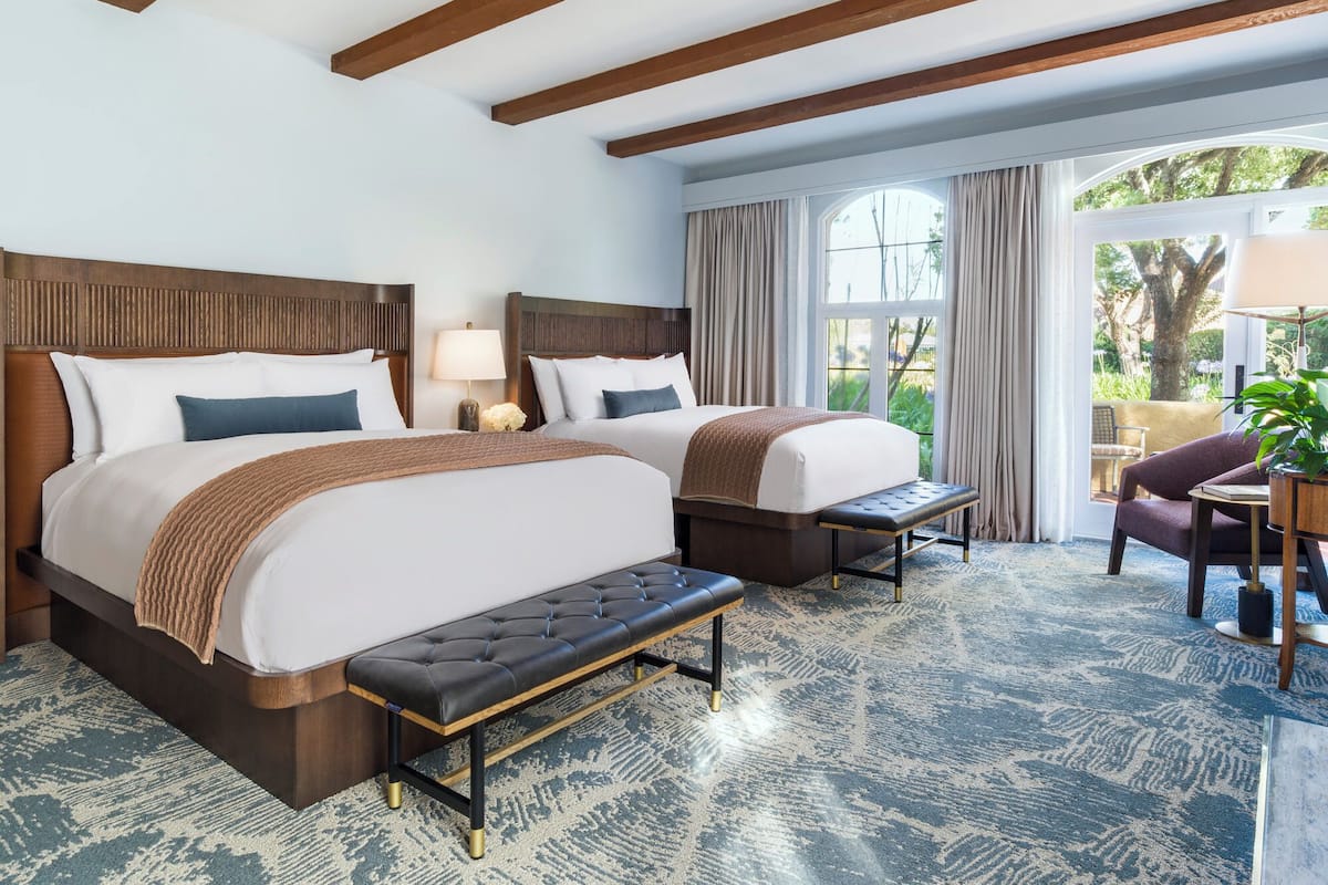 A hotel room with two beds, a bench, a desk, and a view of the outdoors.