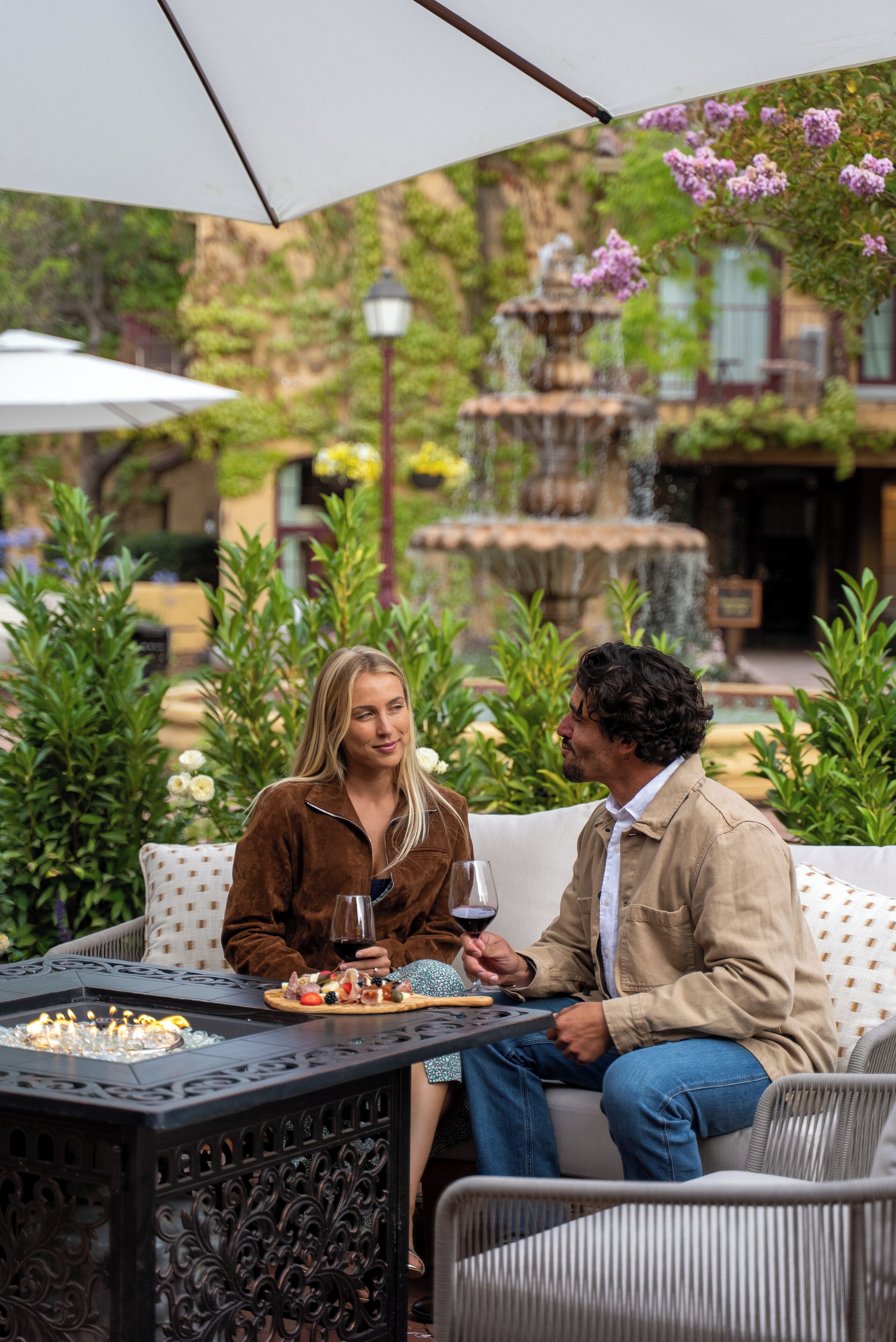 A couple seated at an outdoor table with a decorative fire feature, surrounded by lush greenery and a fountain in the background.