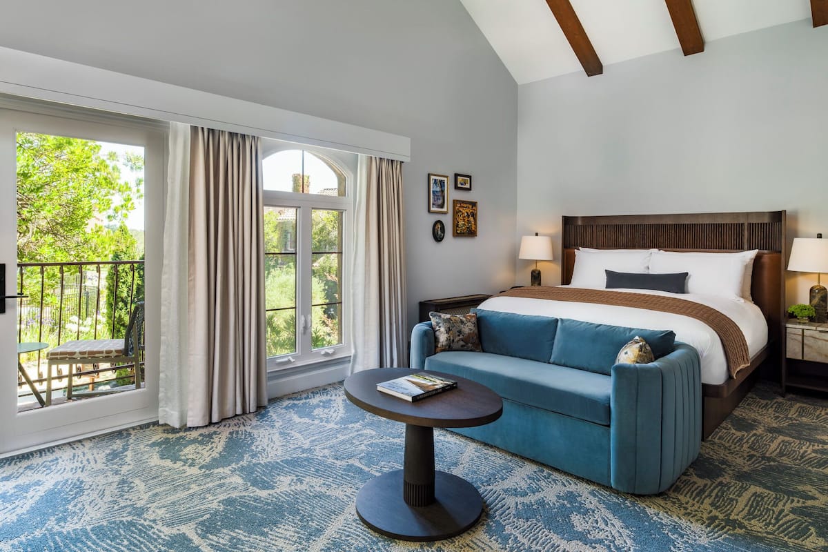 A hotel room with a large window, a bed, a blue sofa, a small round table, and a nightstand with a lamp.