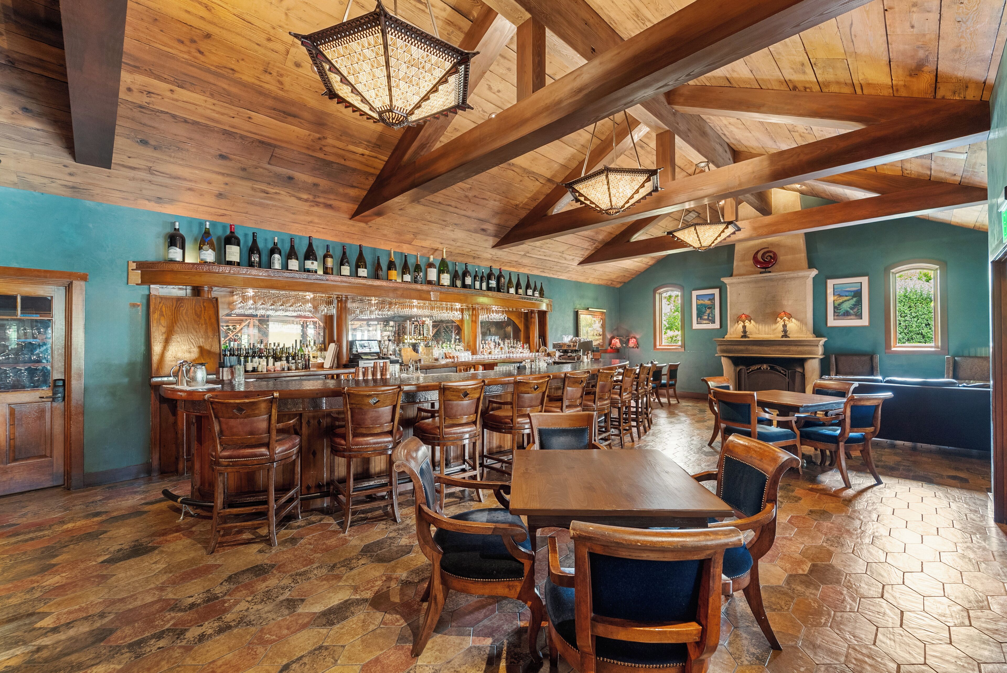 A rustic bar area with wooden furniture, a long counter with wine bottles, and a fireplace.