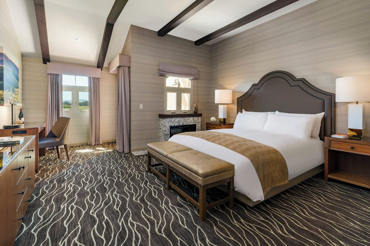 A hotel room with a large bed, a desk, a chair, a bench, and a view of the outdoors.