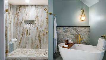 Un baño moderno con una bañera independiente, ducha con cerramiento de vidrio, paredes de mármol y detalles dorados.
