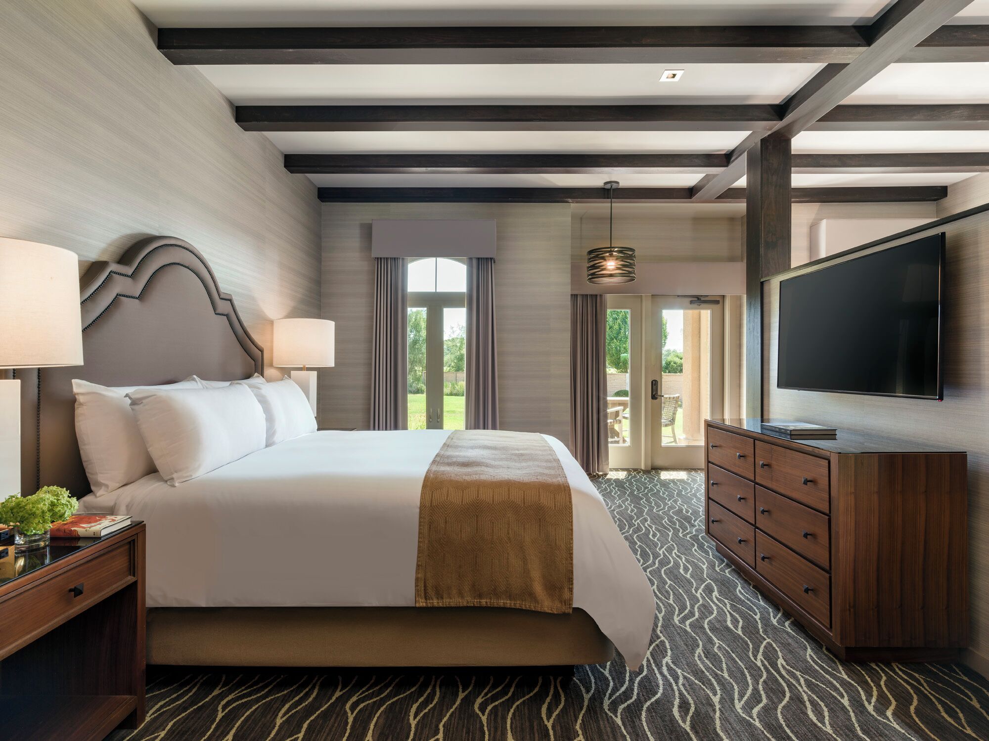 A hotel room with a large bed, a TV, a wooden dresser, and a view of the outdoors.