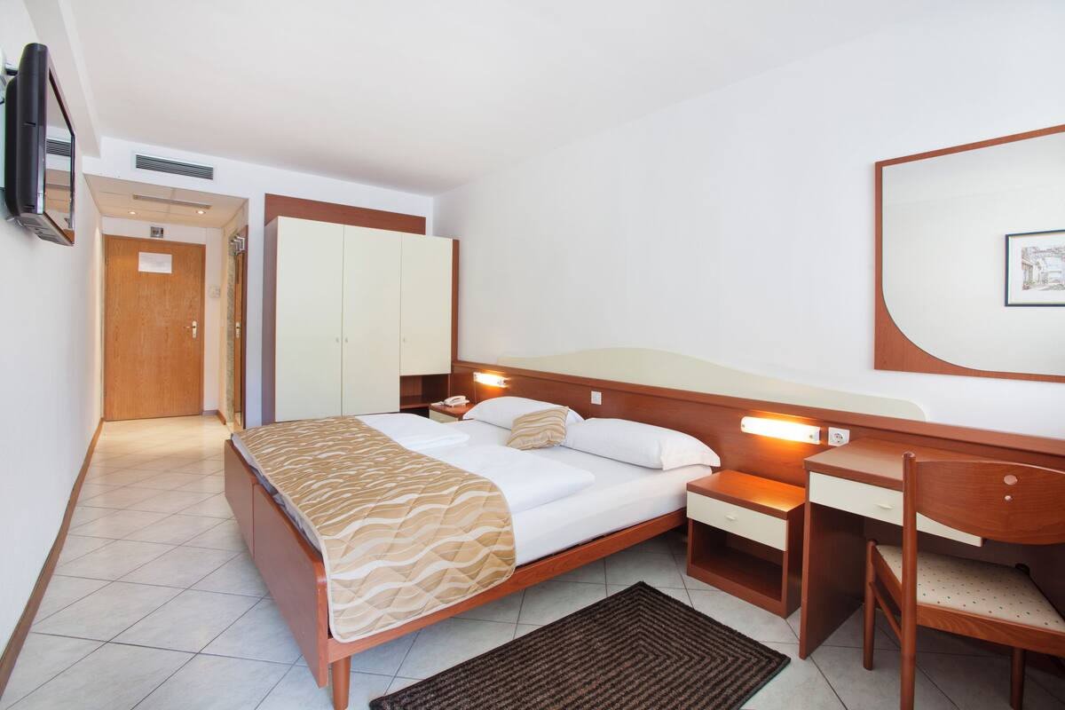 A hotel room with a bed, bedside table, desk, chair, wardrobe, and a television mounted on the wall.