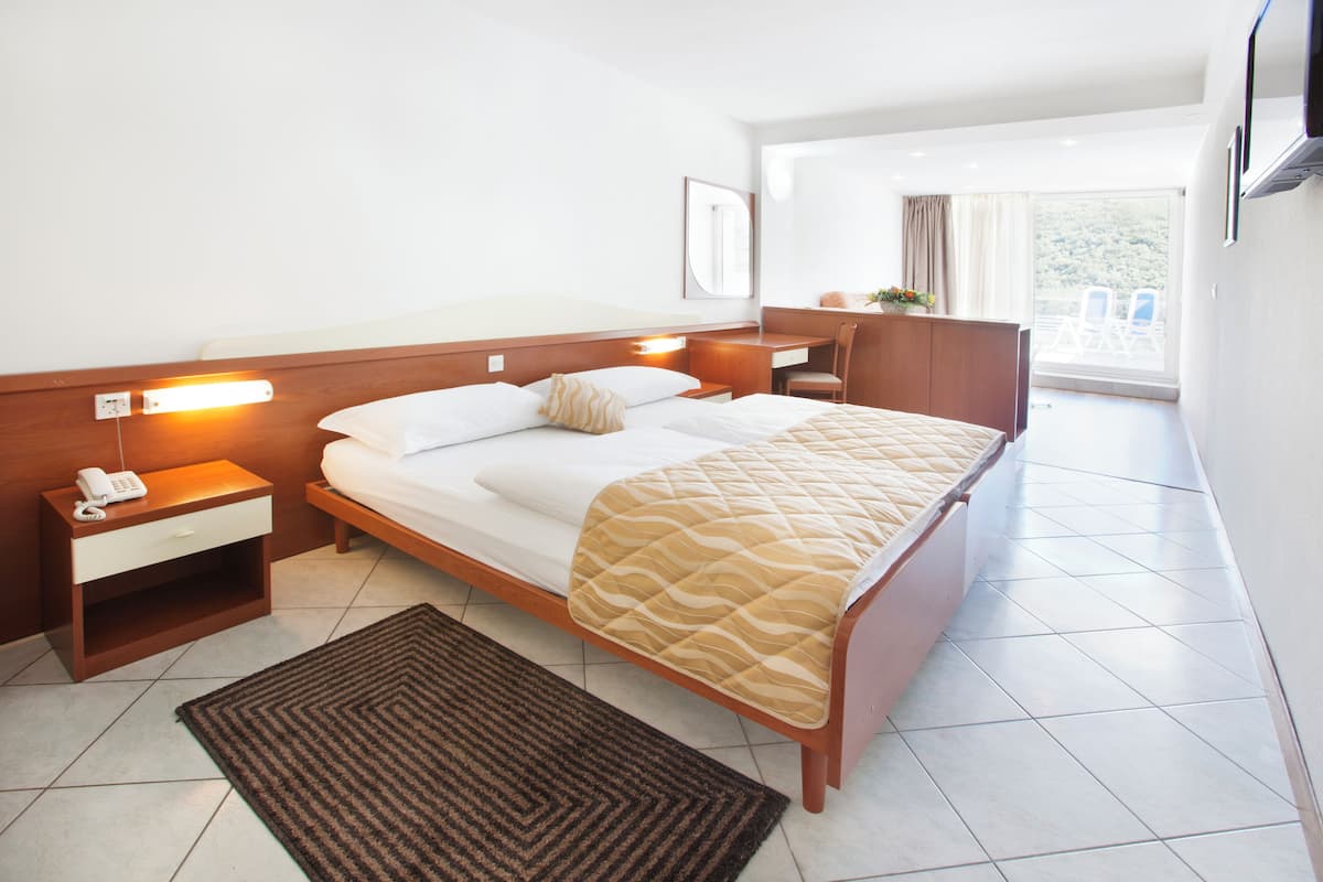 A modern hotel room with a bed, bedside tables, a desk, a chair, and a view of the outdoors.