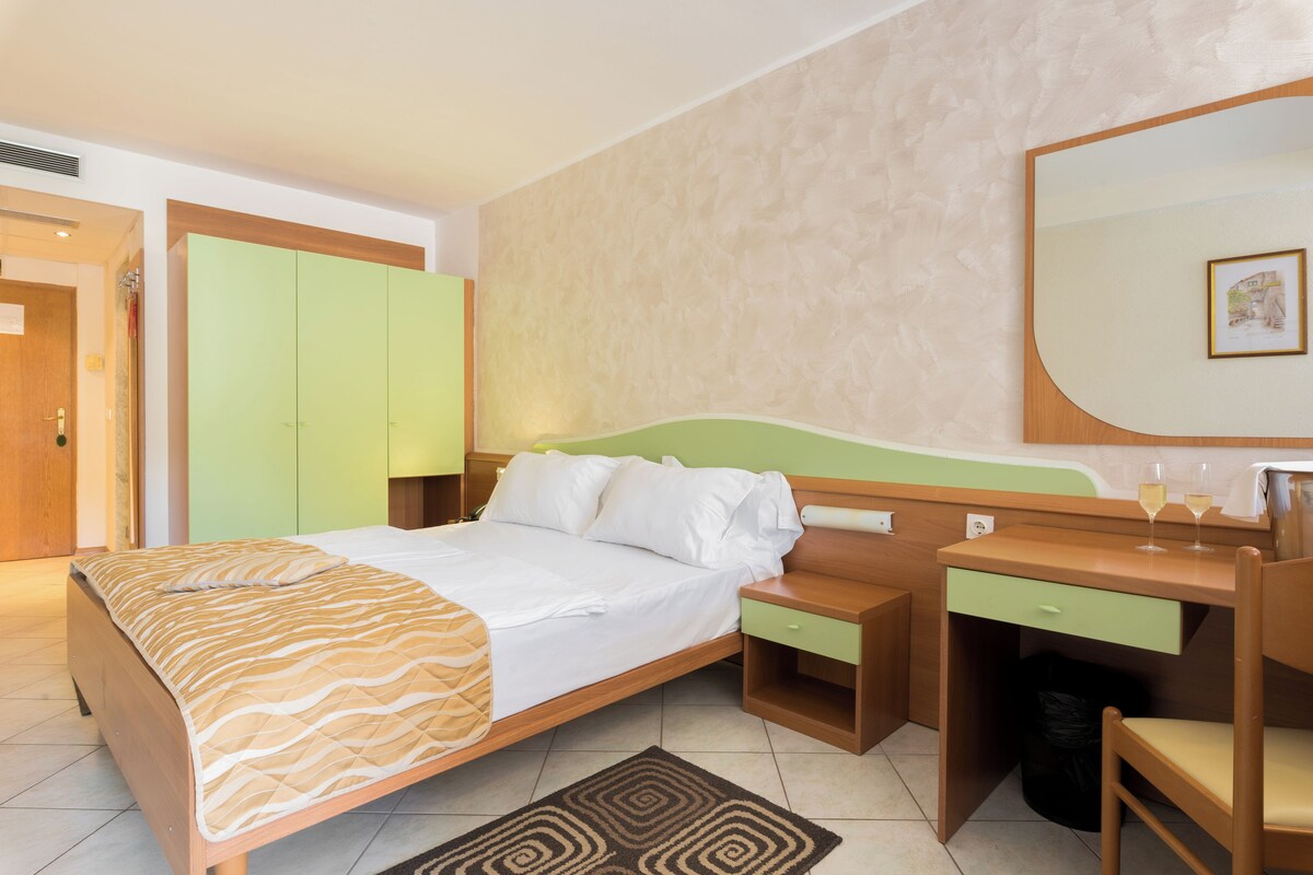 A hotel room with a bed, a desk with a chair, a mirror, and a trash bin.