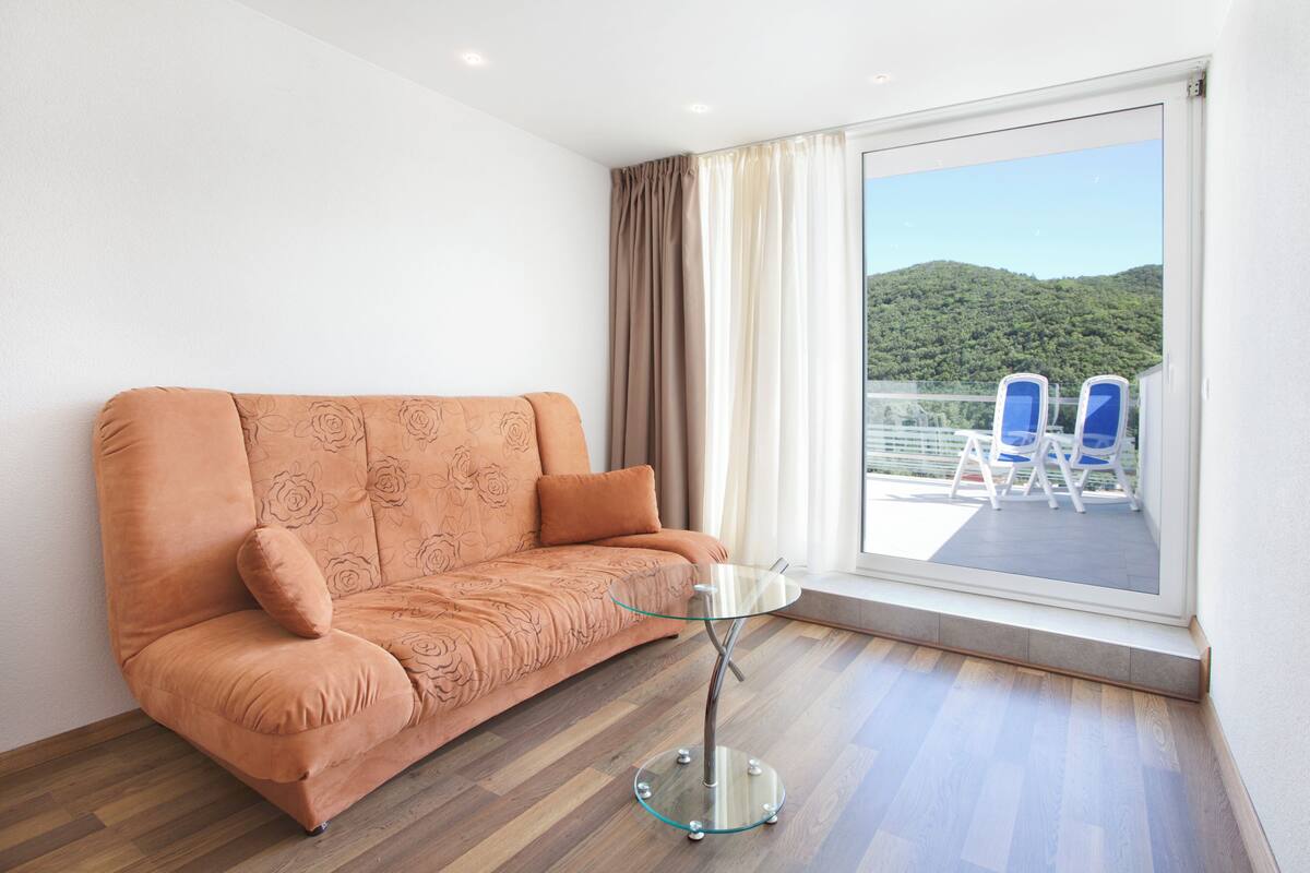A room with a sofa, a glass table, and a view of a balcony with chairs.