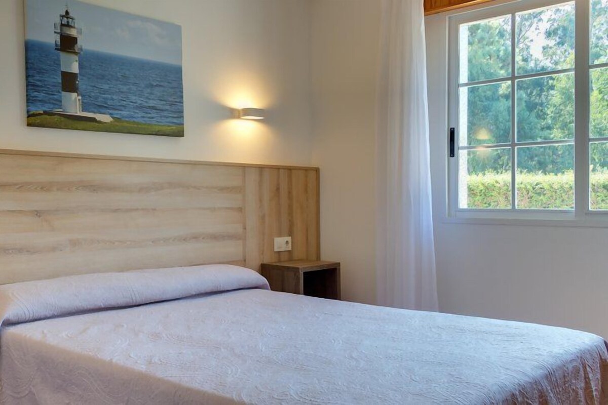A bedroom with a wooden headboard, a painting of a lighthouse, and a window with a view of greenery.