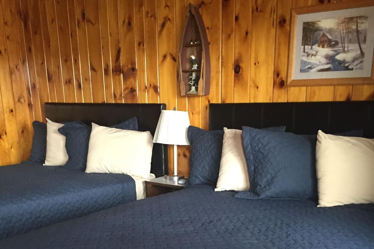 Un lit double avec une couvre-lit matelassée bleue et des oreillers blancs, une table de chevet avec une lampe, et un mur en bois orné d’un tableau encadré.