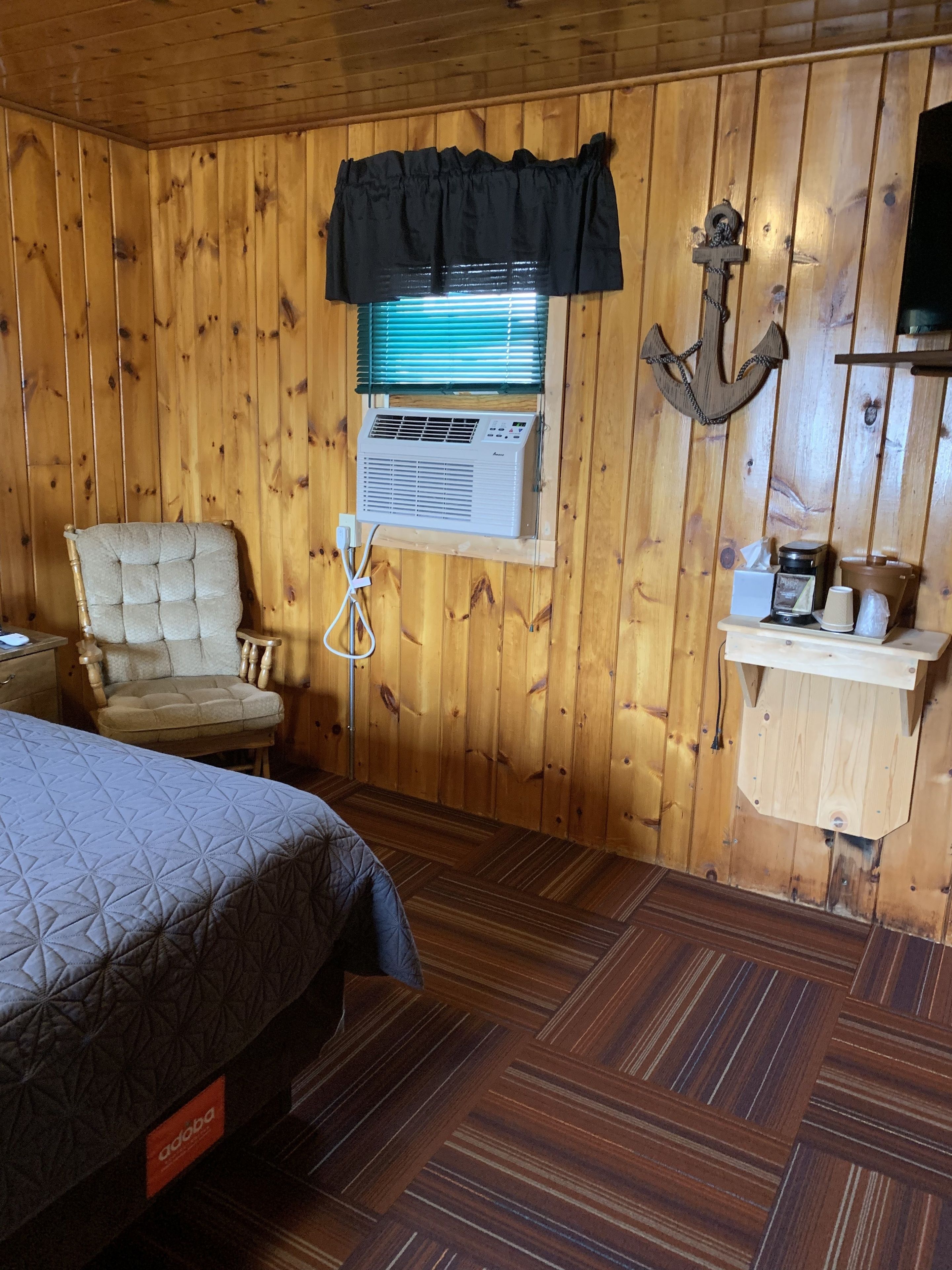 Una habitación con paneles de madera, una cama, una silla, una ventana con persianas, un equipo de aire acondicionado y un pequeño estante con objetos.