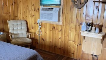 Una habitación con paneles de madera, una cama, una silla, una ventana con persianas, un equipo de aire acondicionado y un pequeño estante con objetos.