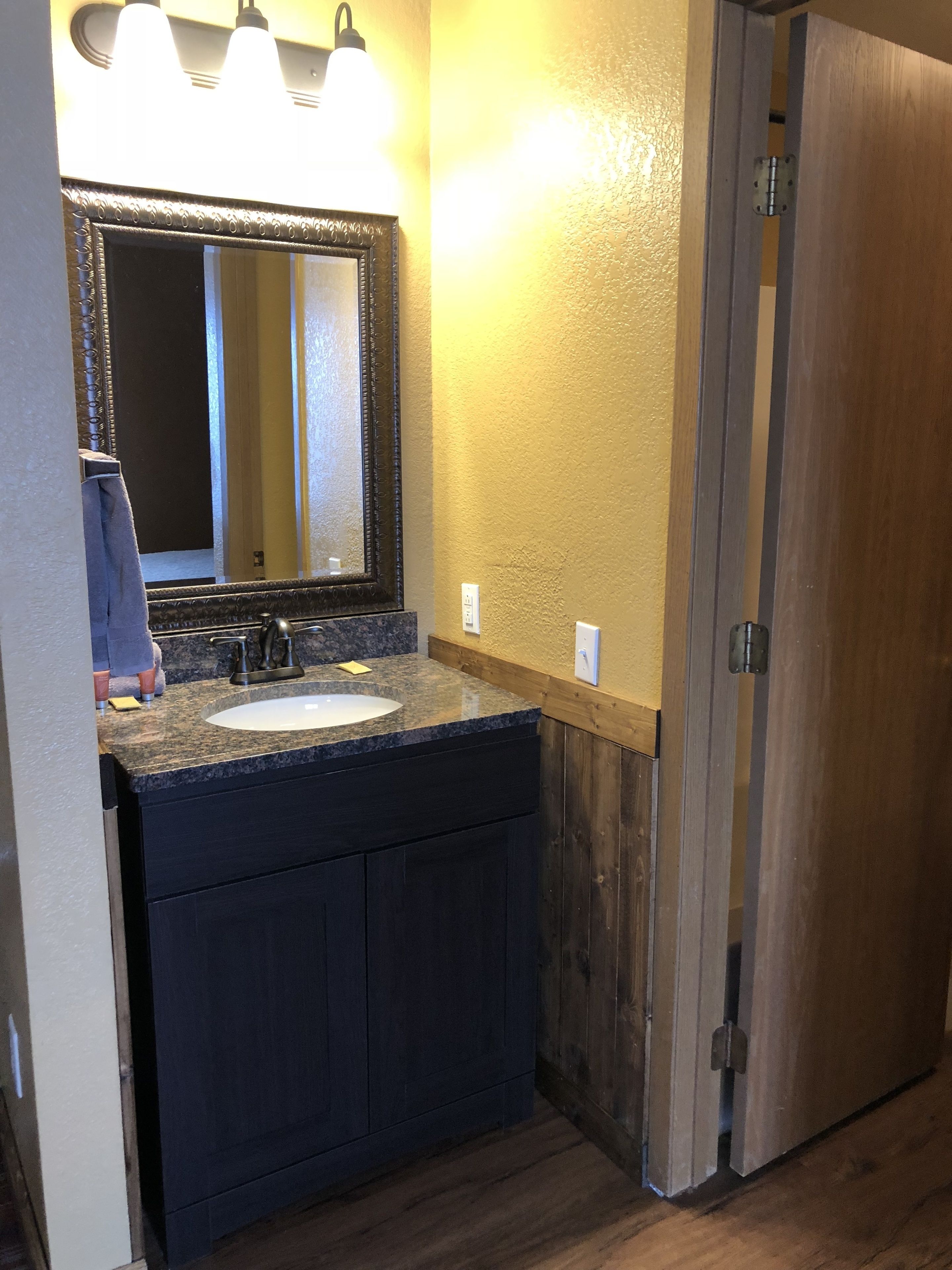 A bathroom with a granite countertop sink, dark cabinets, a large mirror, and wooden walls.