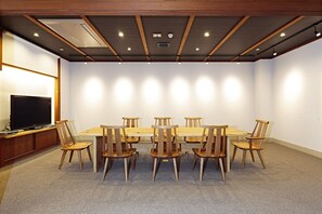 A conference room with a long table, wooden chairs, a television on a stand, and recessed ceiling lighting.