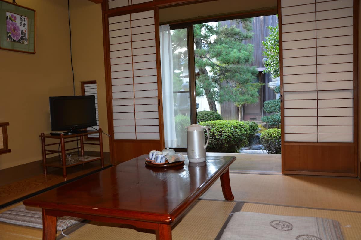 A traditional Japanese room with tatami flooring, a television on a wooden stand, and a view of a garden through shoji screens.