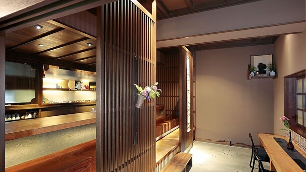 A traditional Japanese-style interior with wooden sliding doors, a dining area with a wooden table and chairs, and a kitchen visible through the sliding doors. - Katsuzakikan (Tsubata)