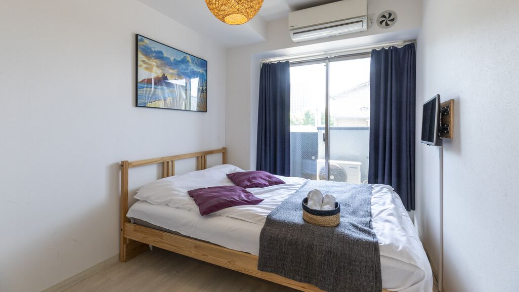 Un lit simple avec un cadre en bois, une literie blanche et des oreillers violets. Il y a une fenêtre avec des rideaux bleus, un climatiseur fixé au mur et un tableau au-dessus du lit.