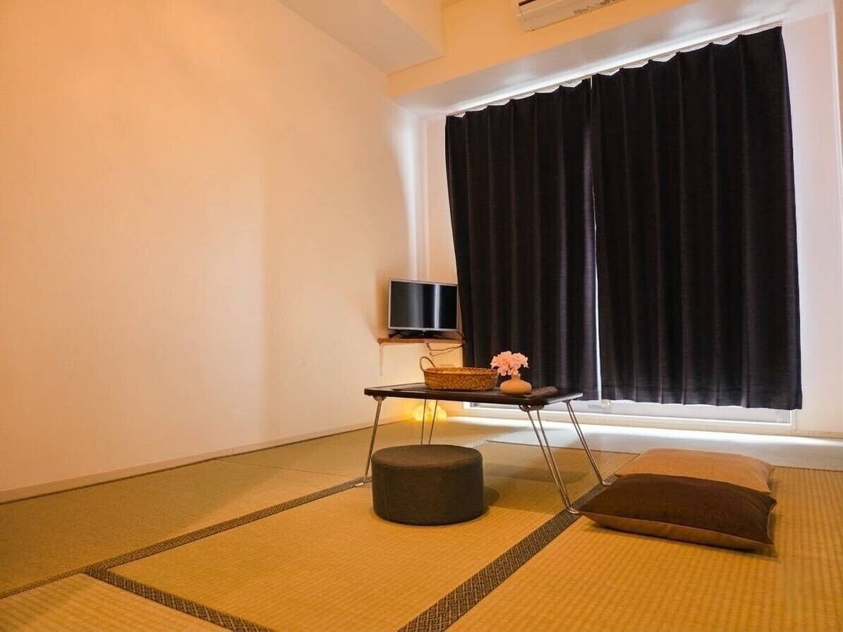 Une pièce avec un sol recouvert de tatamis, un téléviseur noir posé sur un meuble, une petite table avec un panier et un vase, et une fenêtre avec des rideaux noirs.