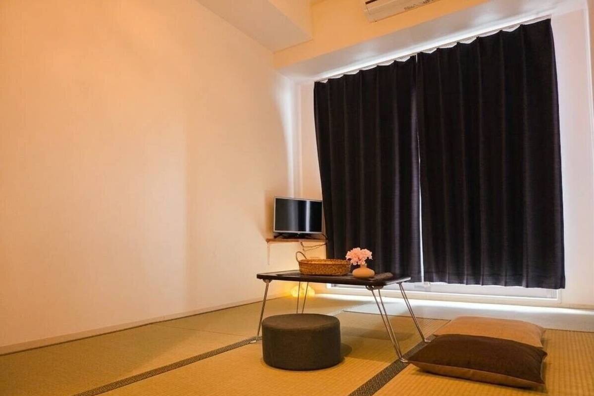 Ett rum med tatamimattor på golvet, en svart tv på ett tv-bord, ett litet bord med en korg och en vas, och ett fönster med svarta gardiner.