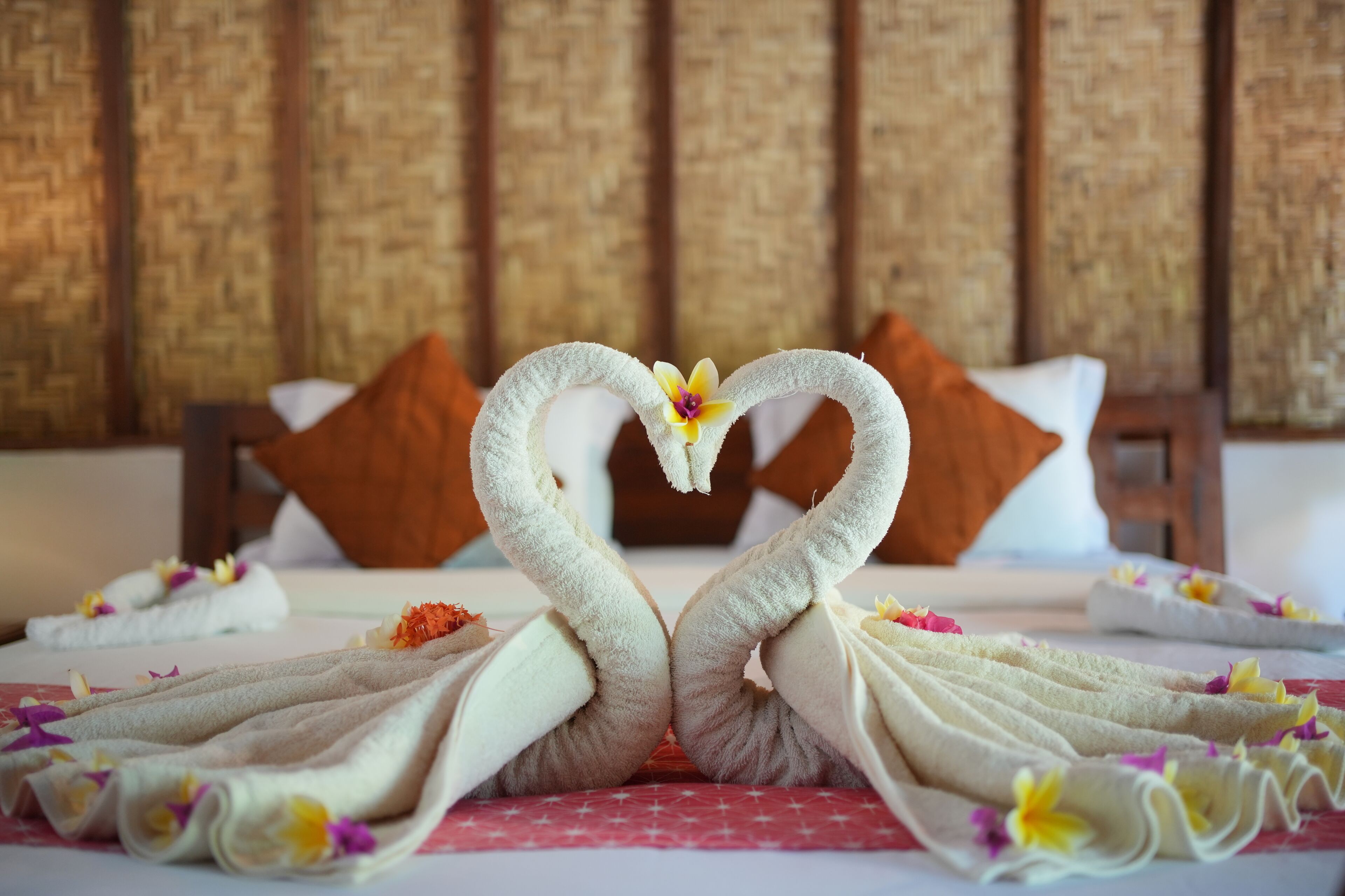 Cama com cabeceira trançada, almofadas brancas e arranjo de toalhas decorativo em formato de cisnes com flores.