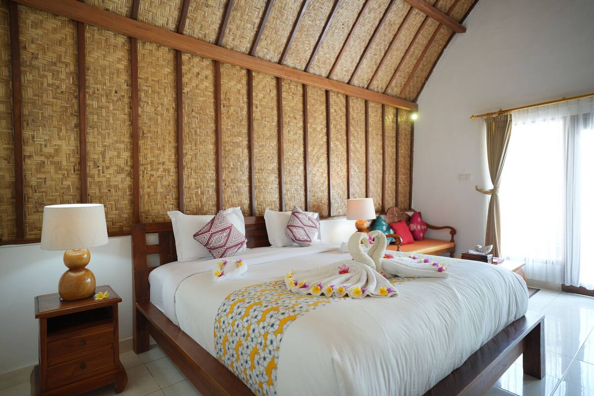 A neatly made bed with a white comforter and decorative swan pillows, a wooden nightstand with a lamp, and a woven bamboo ceiling.