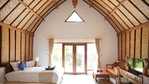 Una habitación amplia con techo alto de paja, una cama grande, un escritorio con televisor y un puerta corrediza de vidrio que da a un área exterior.