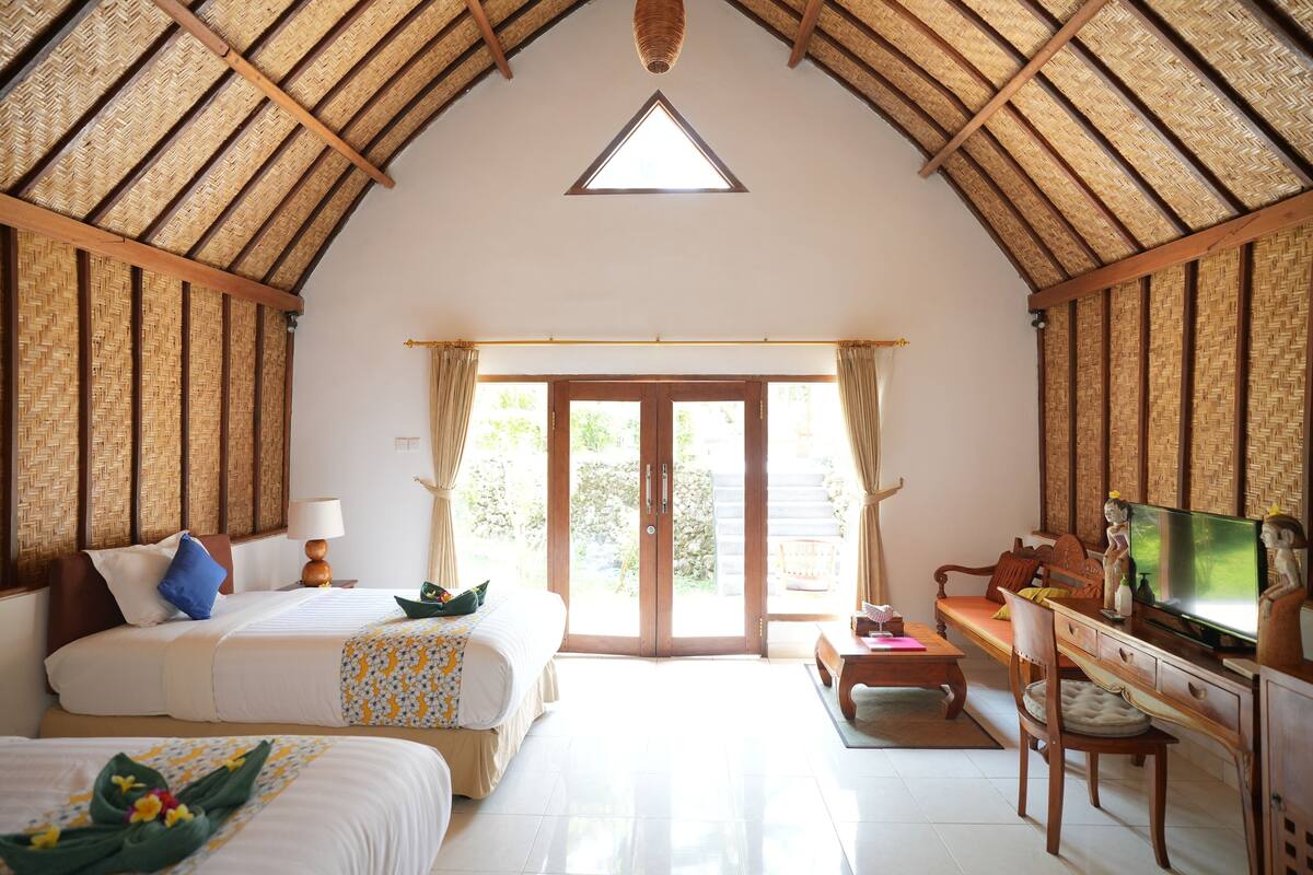 A spacious room with a high, thatched ceiling, a large bed, a desk with a television, and a sliding glass door leading to an outdoor area.
