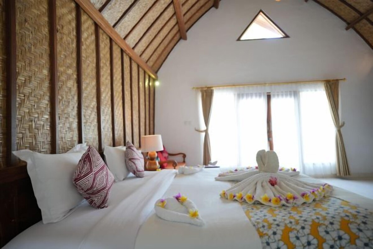 Ett hotellrum med högt tak, en vägg med vävt mönster, en säng med vita lakan och ett blommigt överkast, en sänglampa och ett fönster med gardiner.