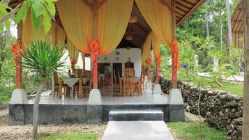 A covered outdoor seating area with a table and chairs, surrounded by greenery and a stone wall.