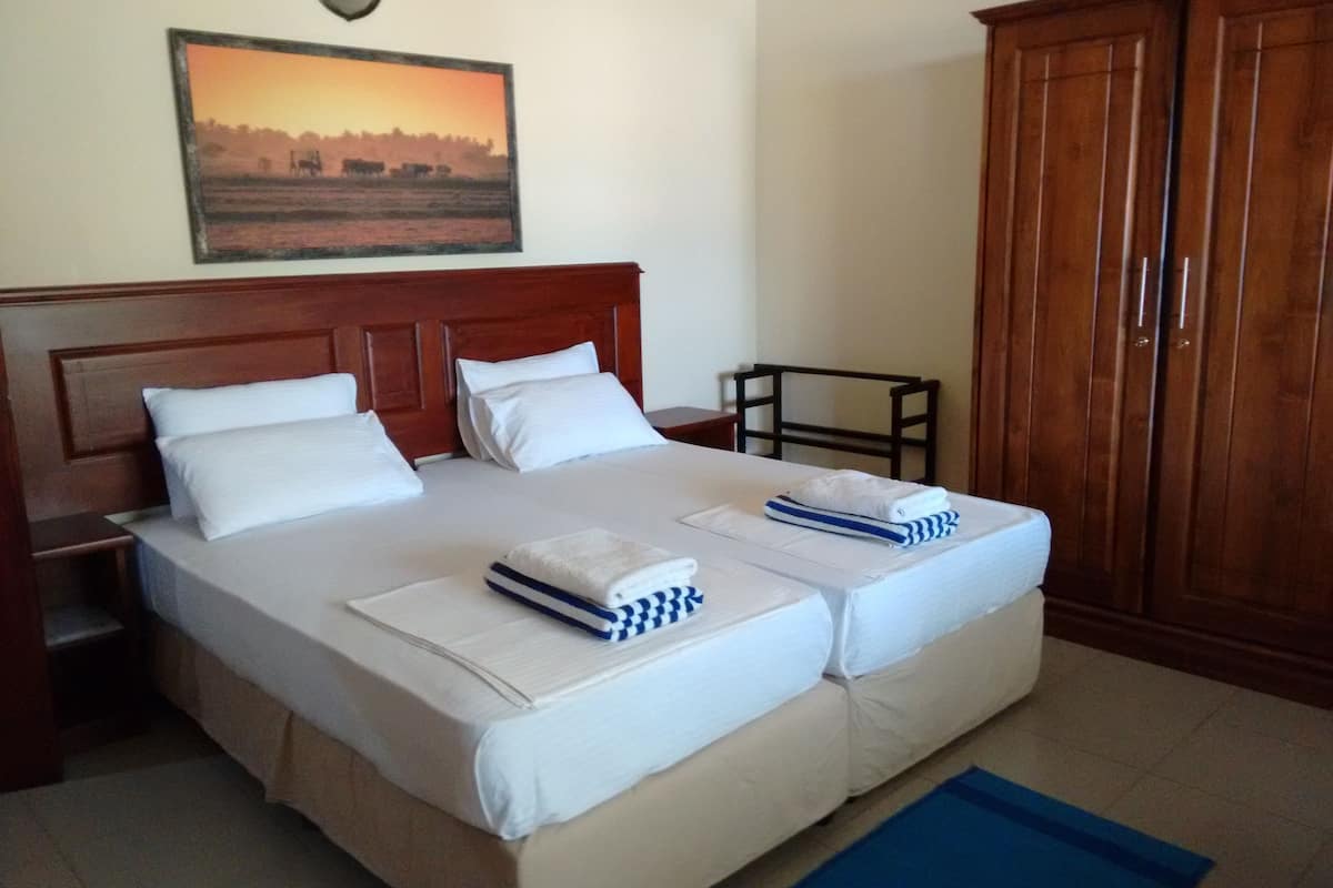 A double bed with white linens, a wooden headboard, and a blue and white striped towel. There is a wooden wardrobe to the right and a framed picture on the wall above the bed.