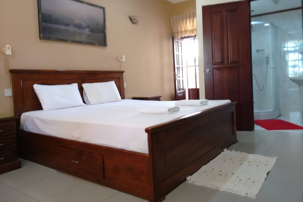 A bedroom with a wooden bed, a nightstand, a wall-mounted light, a window with curtains, and a door leading to a bathroom.