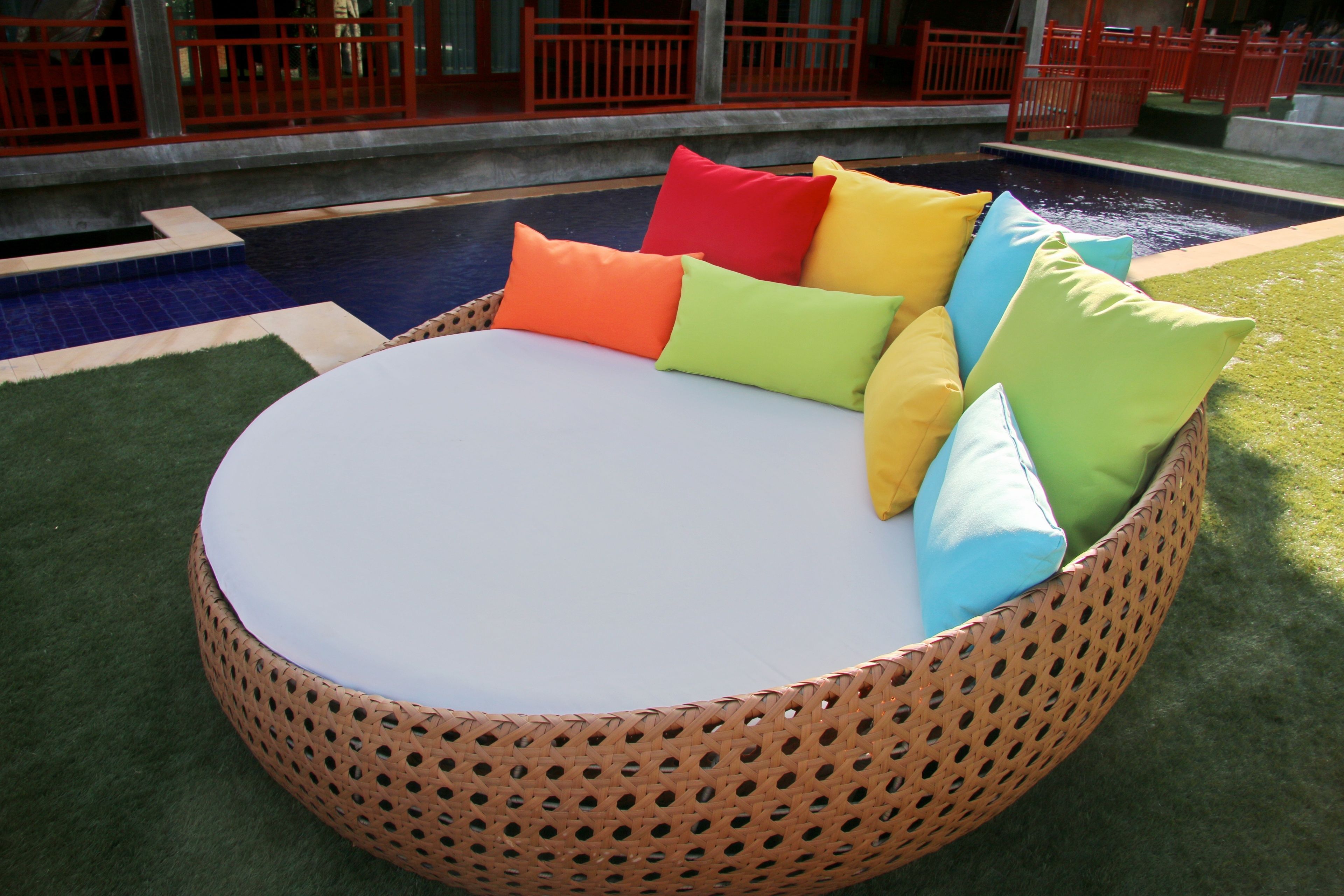 A round wicker lounge chair with a white cushion and colorful cushions, placed on artificial grass near a pool.