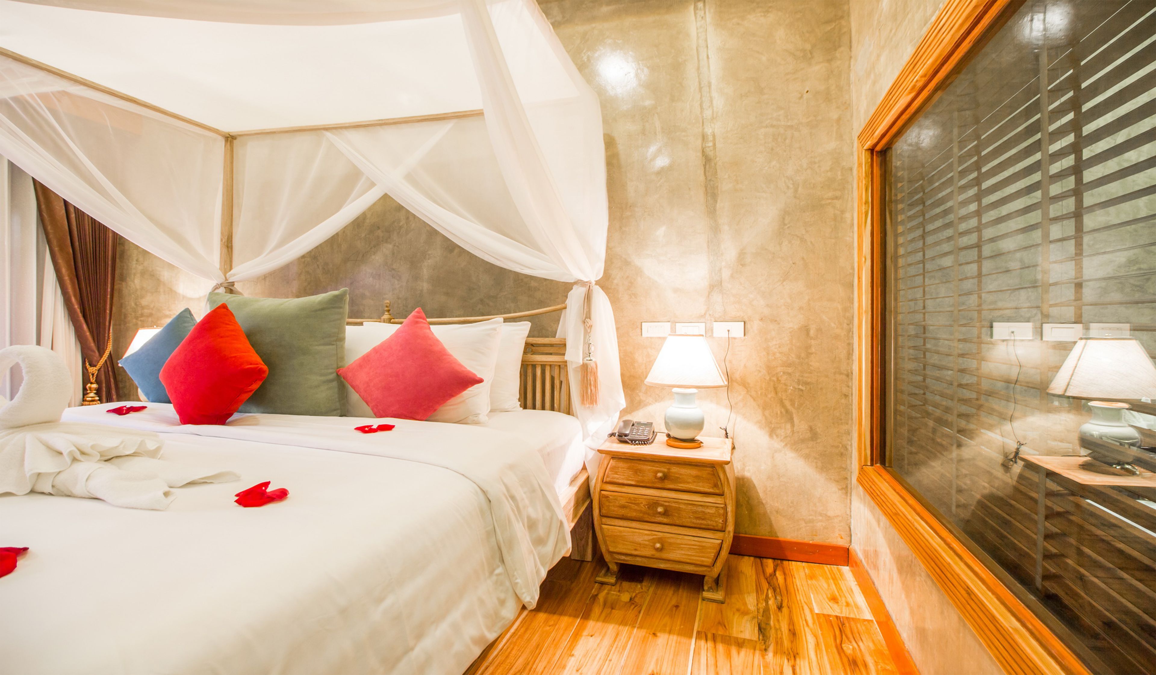 Habitación de hotel con cama con dosel, mesita de noche con lámpara y un gran ventanal con persianas.