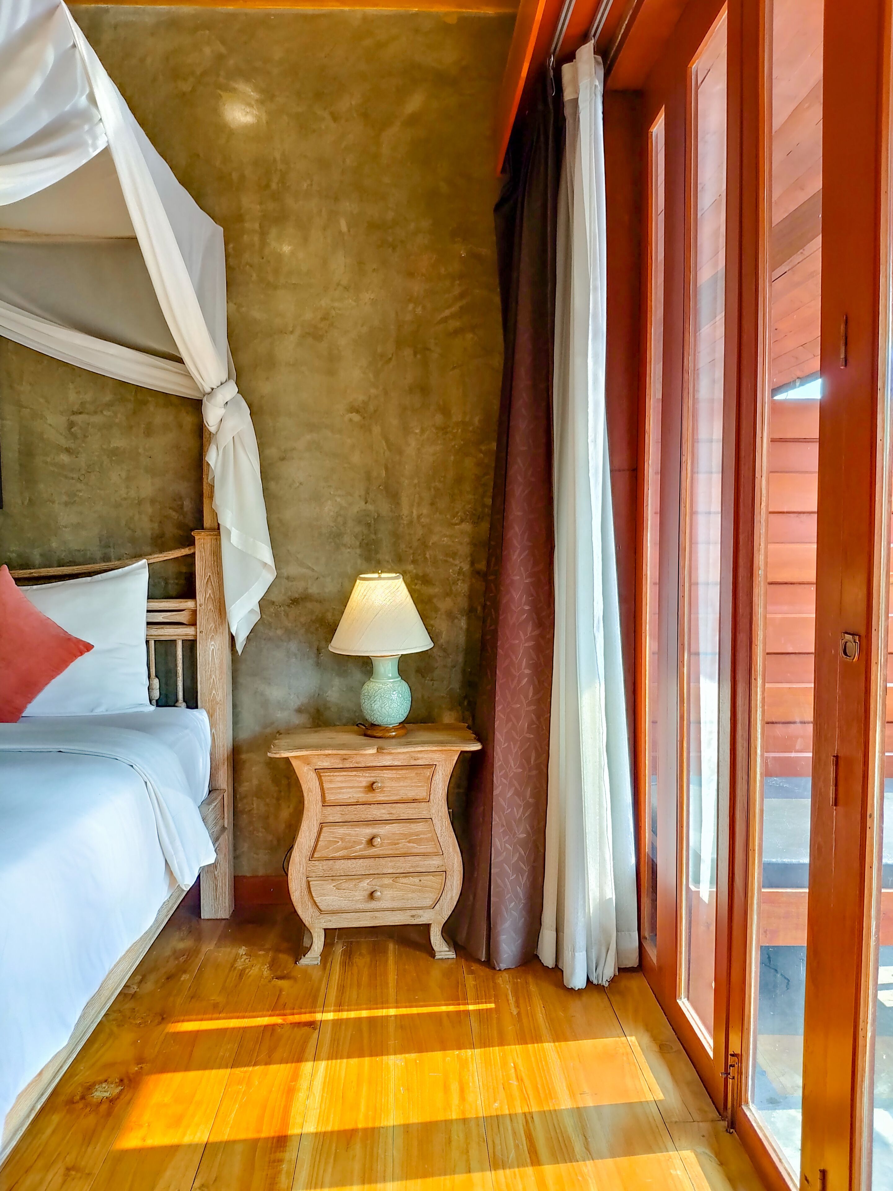 A bedroom with a wooden floor, a bed with a canopy, a nightstand with a lamp, and a sliding glass door.